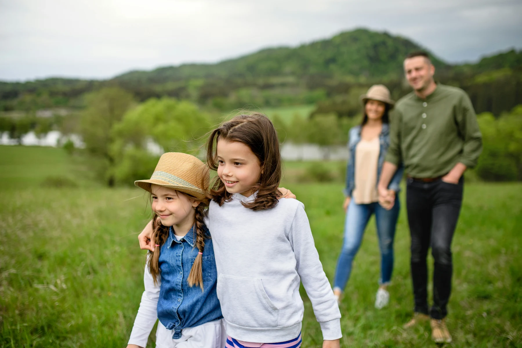Two sisters standing together with their parents nearby in a green field.