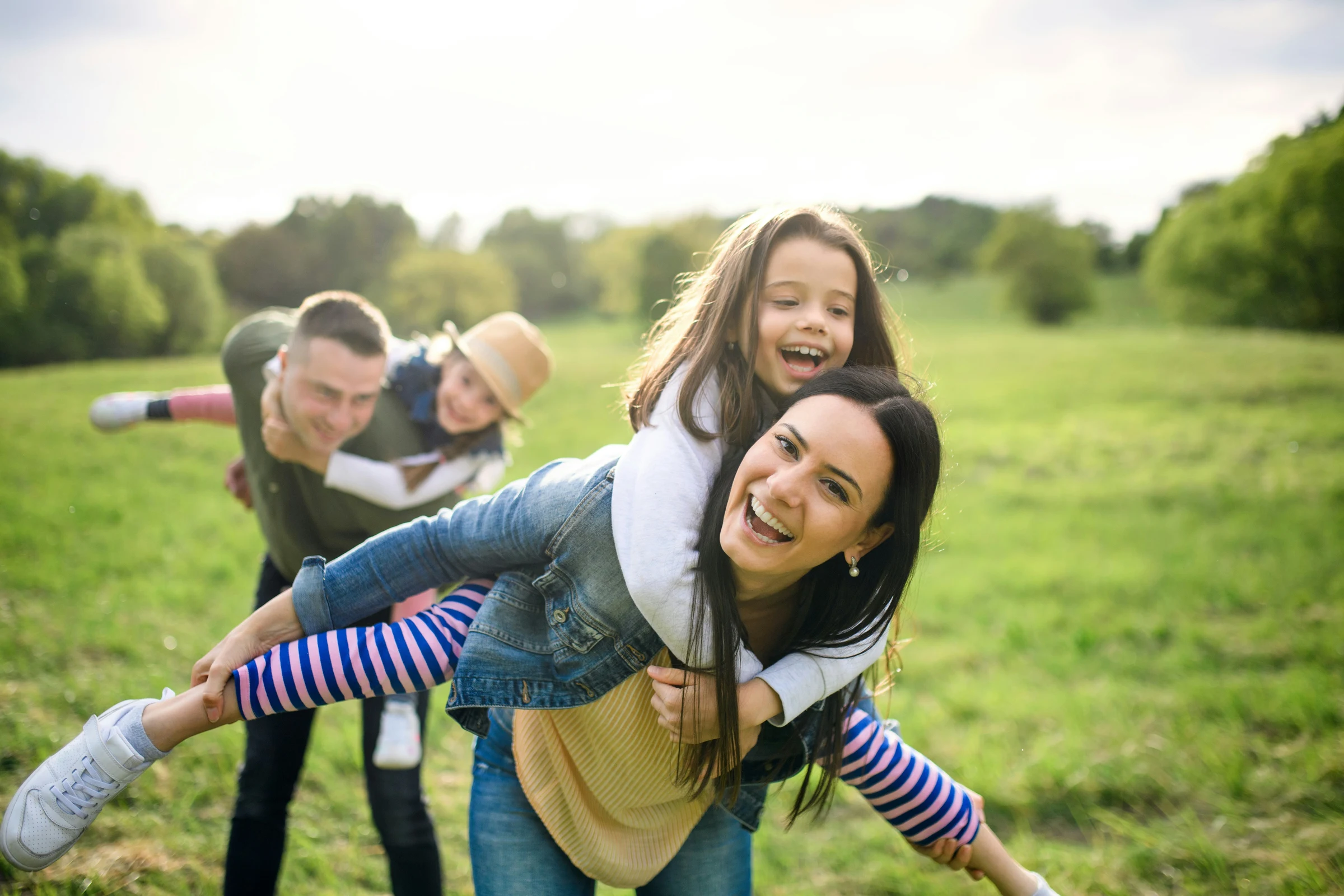 Parents giving their two daughters piggyback rides together in a sunny field.